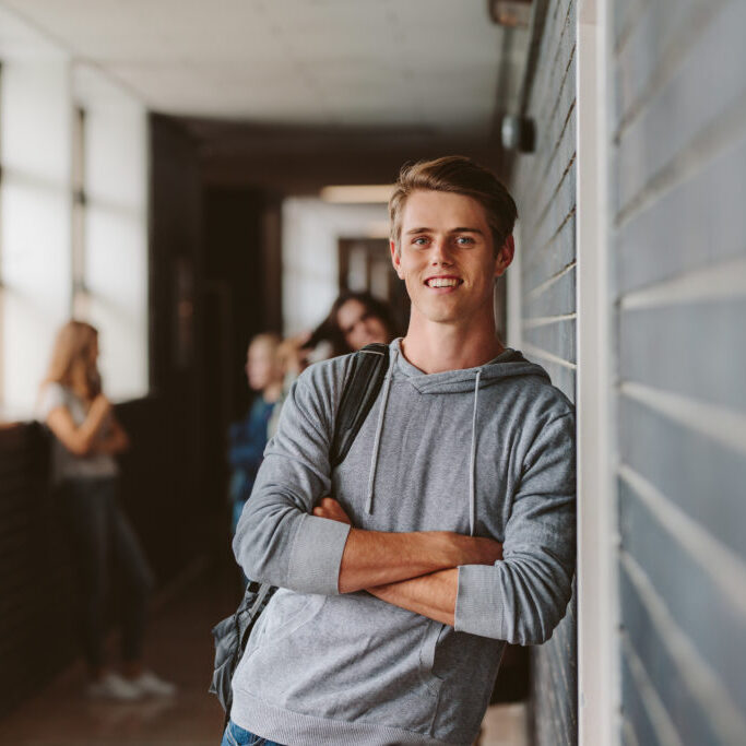Portrait of handsome young university student standing and leaning to a wall in college corridor with other students at the back. Caucasian male student in high school campus.